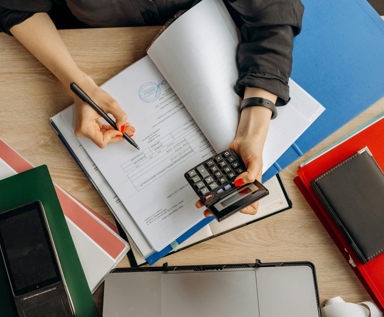 A person with a calculator in hand works on documents and contracts at a desk full of papers.