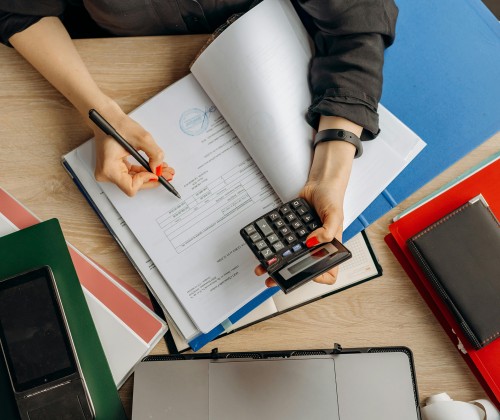 A person with a calculator in hand works on documents and contracts at a desk full of papers.
